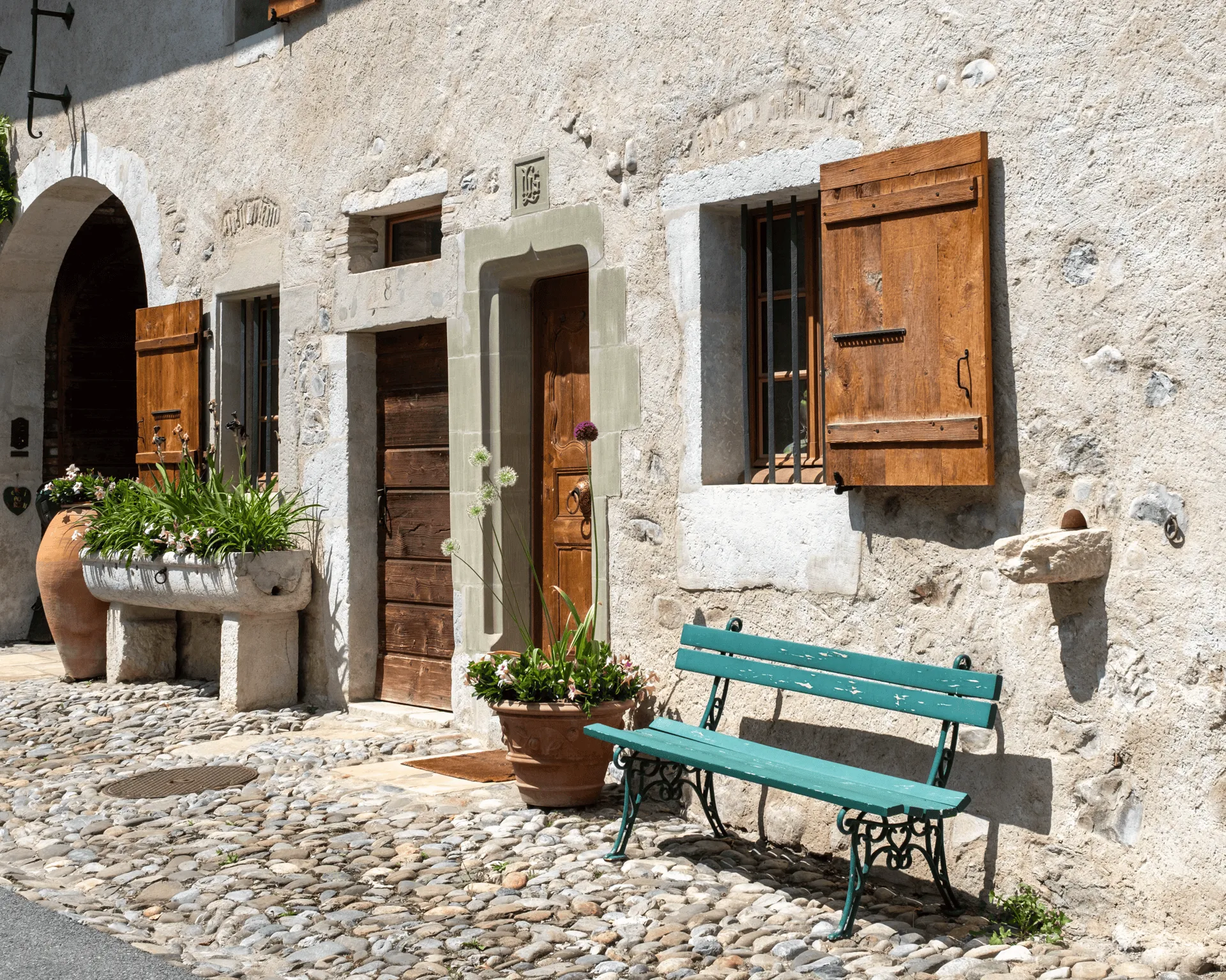 "A wooden bench painted teal blue. Behind it is a brightly lit white building with wooden doors and windows "