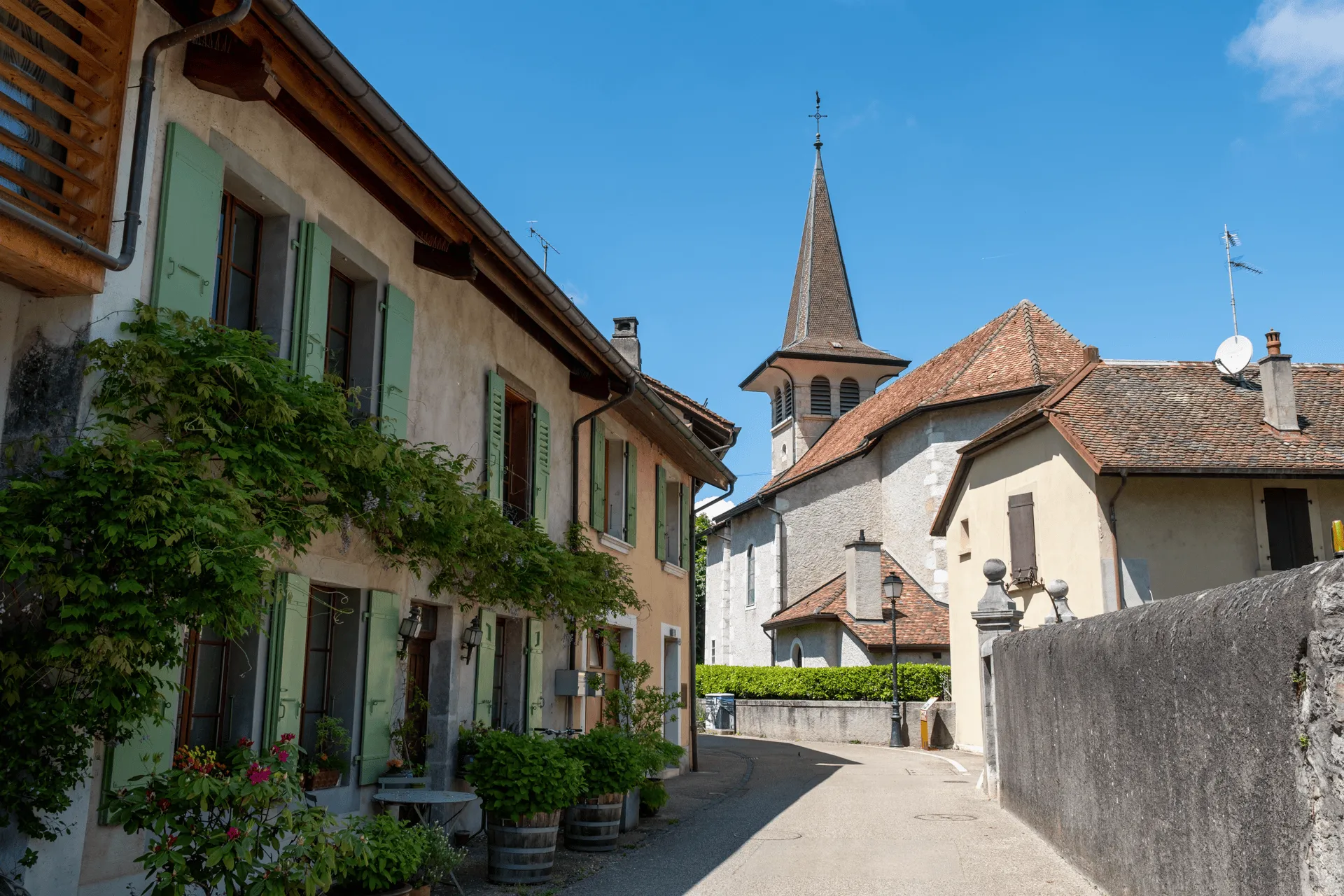 "A view of the lane leading through the Swiss village of Veyrier. You can see green-painted window doors and a church in the distance."